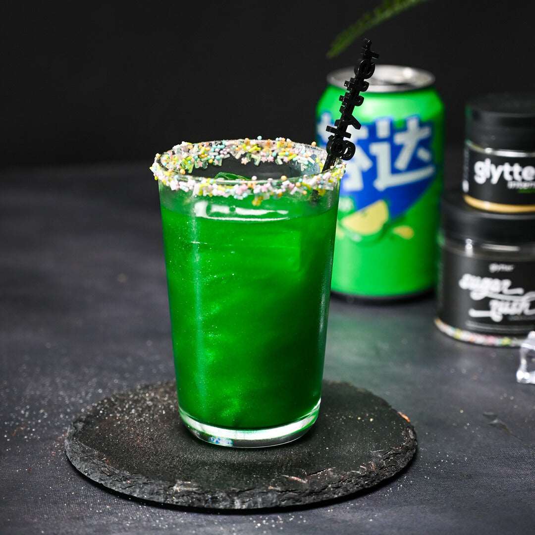 Decocino Glytter Intense Jungle Green – grünes glitzerndes Getränk im Glas mit buntem Zuckerrand und Produktdosen im Hintergrund