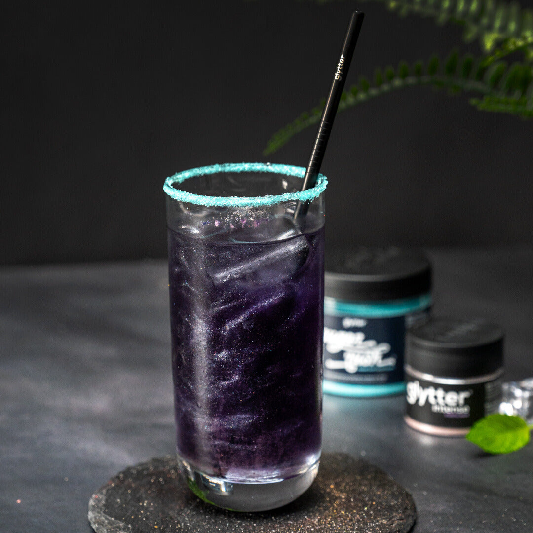 Glytter Intense Purple Rain – violettes glitzerndes Getränk im Glas mit türkisfarbenem Zuckerrand und Produktdosen im Hintergrund
