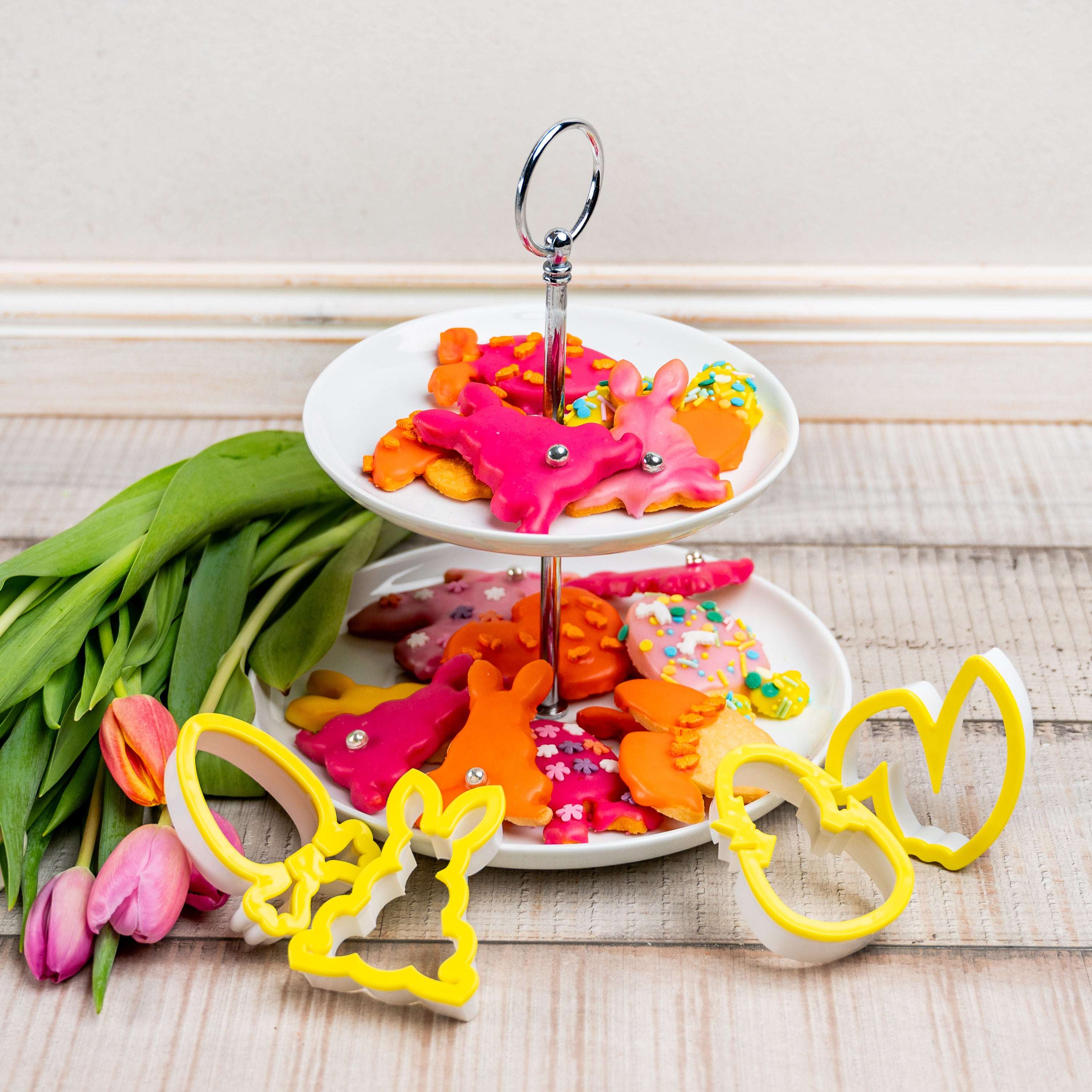 Osterkekse in bunten Farben auf einer Etagere, dekoriert mit gelben Deko Back Ausstechern in Hasen- und Ostereiformen