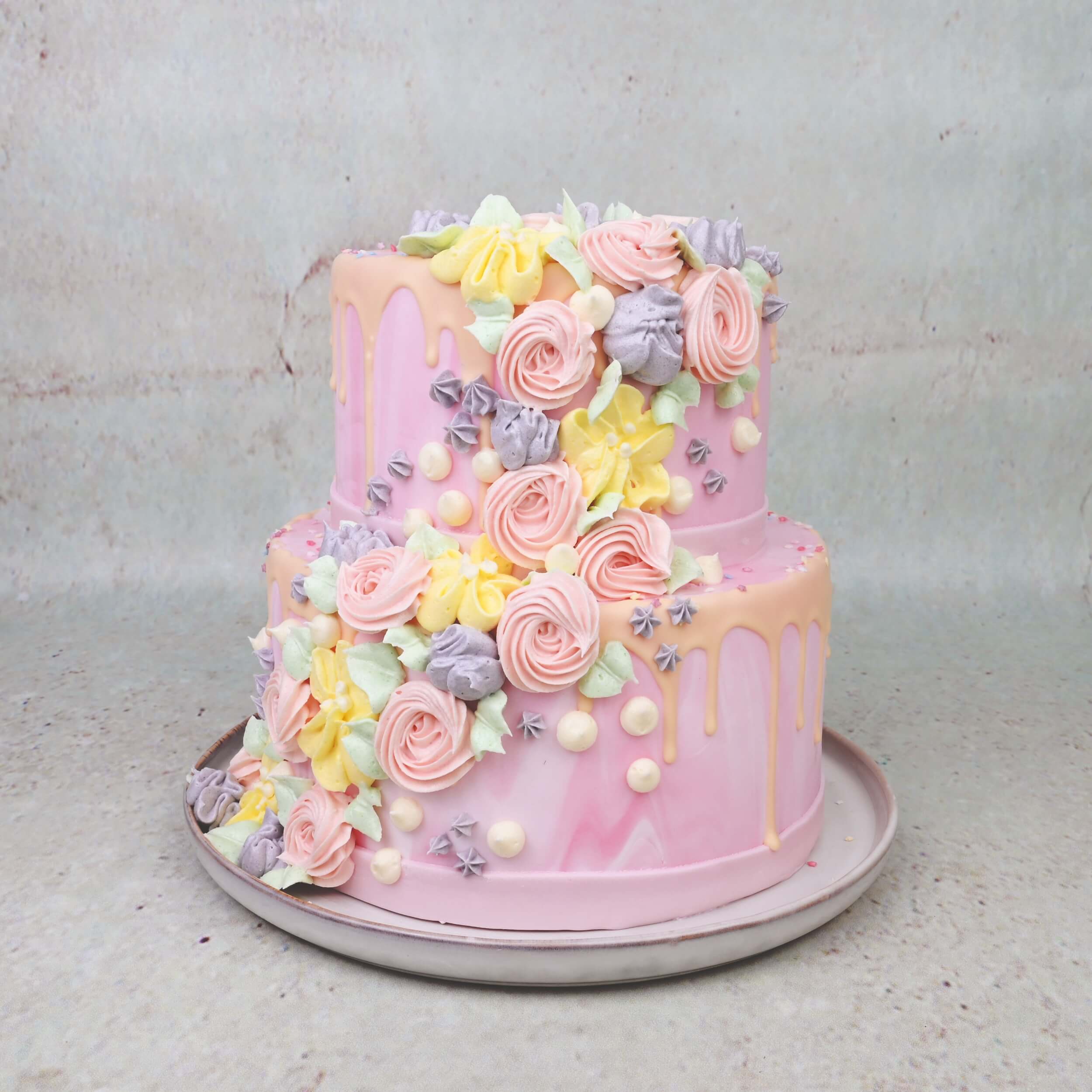 Zweistöckige rosa Torte mit pastellfarbenen Buttercreme-Blumen und Drip-Dekor, dekoriert mit Decocino Spritztüllen