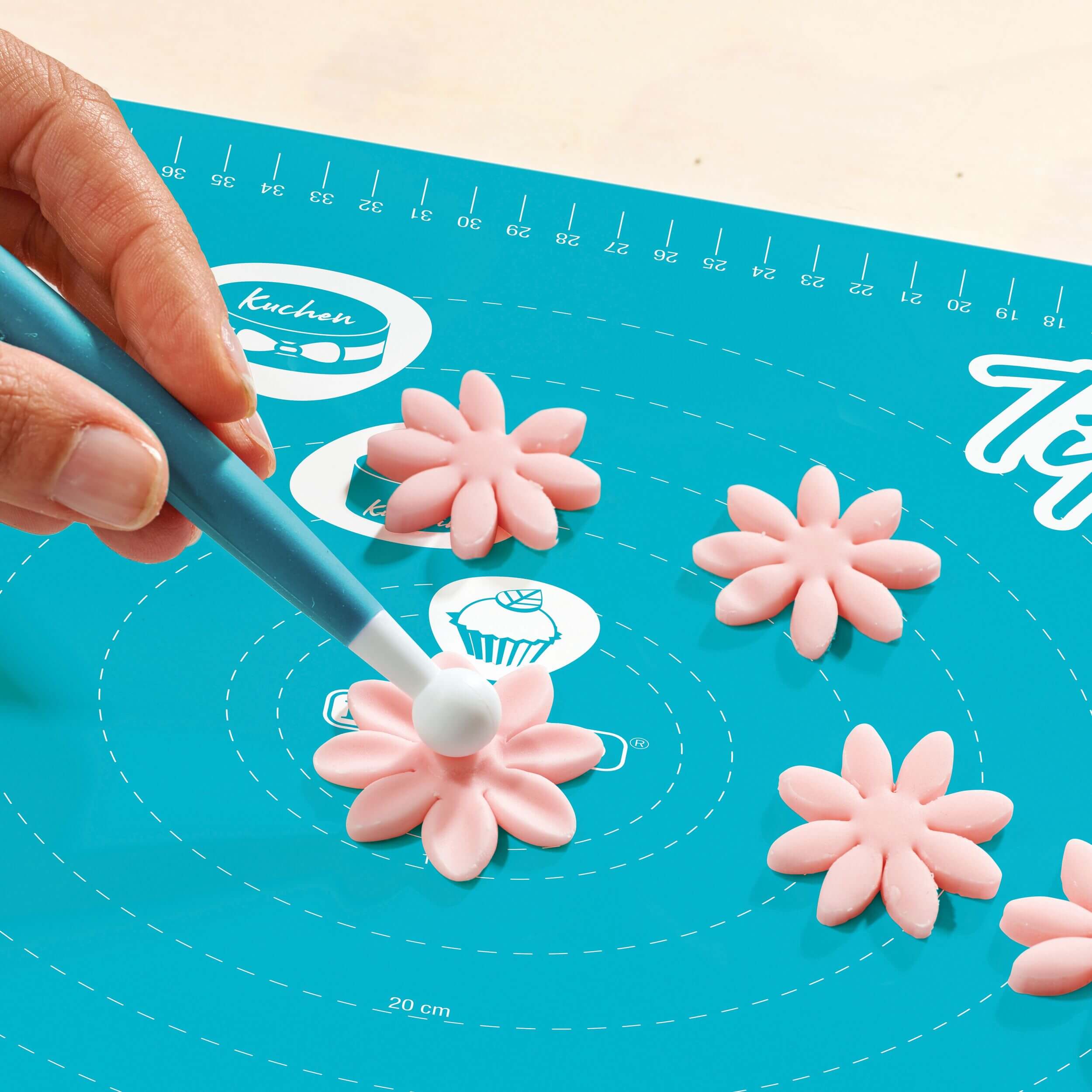 Hand formt rosa Fondantblume mit einem Modellierwerkzeug auf einer türkisfarbenen Decocino Back- und Ausrollmatte