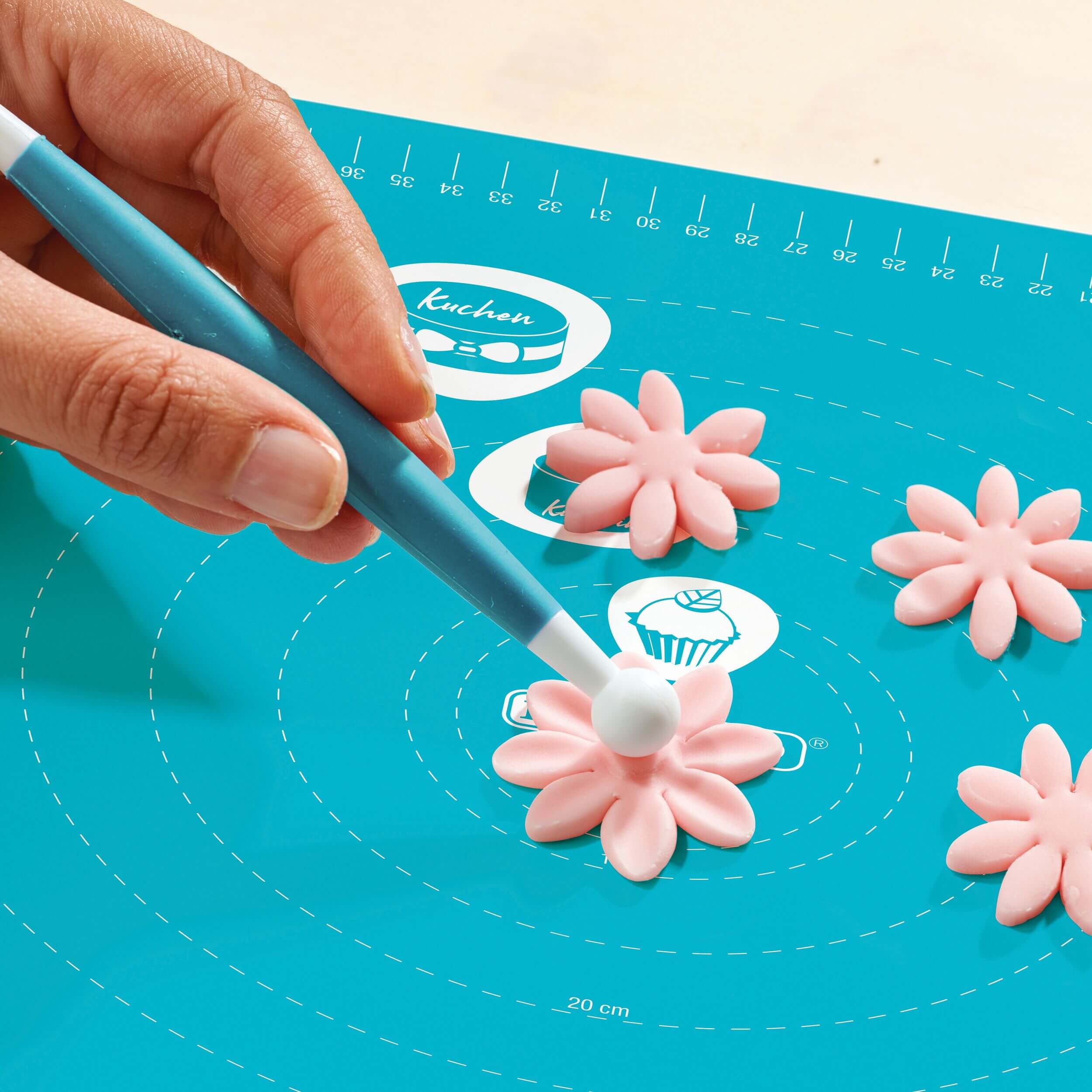 Hand formt rosa Fondantblume mit einem Modellierwerkzeug auf einer türkisfarbenen Decocino Back- und Ausrollmatte