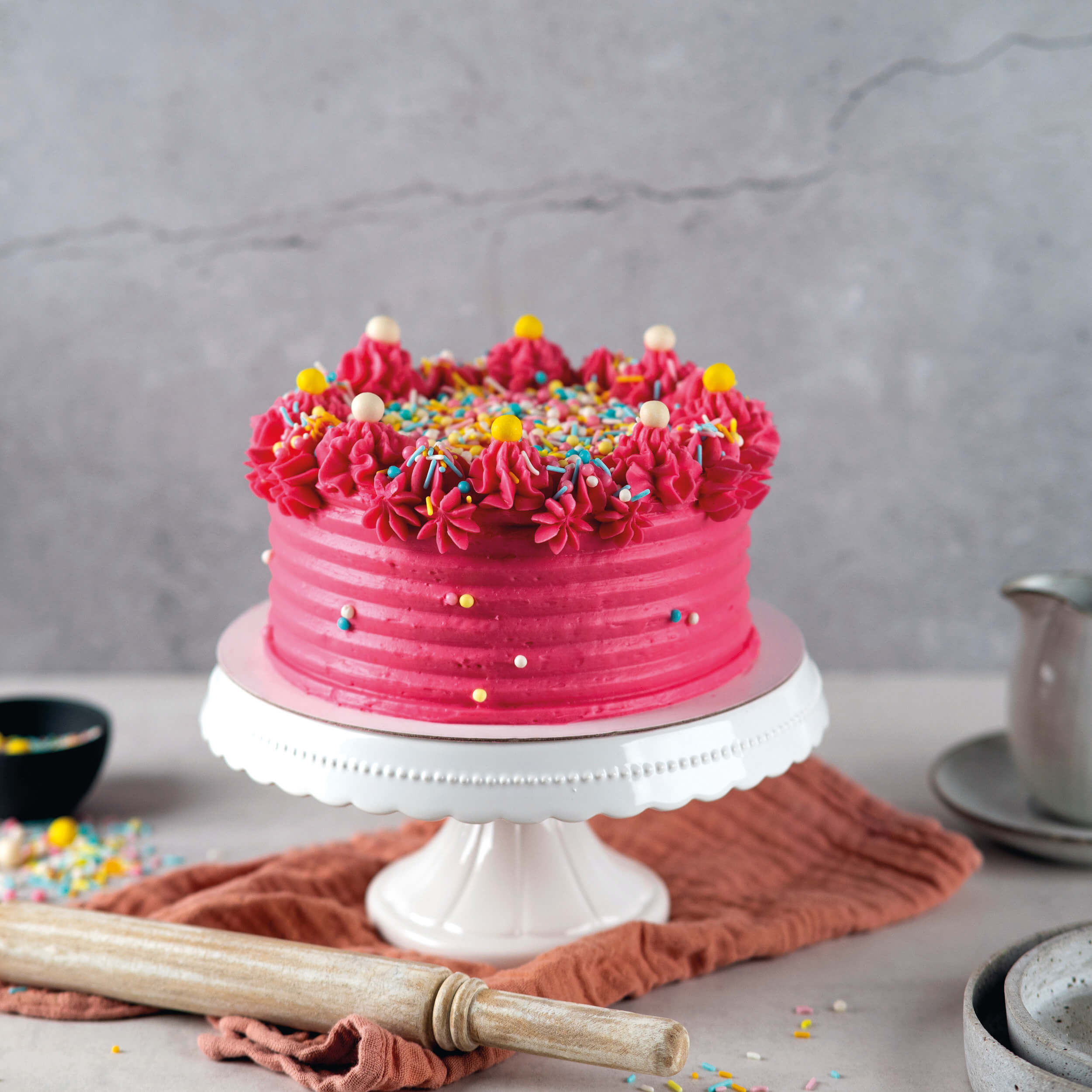 Pink eingestrichene Torte mit Cremetuffs und bunten Streuseln auf Tortenständer – dekoriert mit Decocino Sprinkles