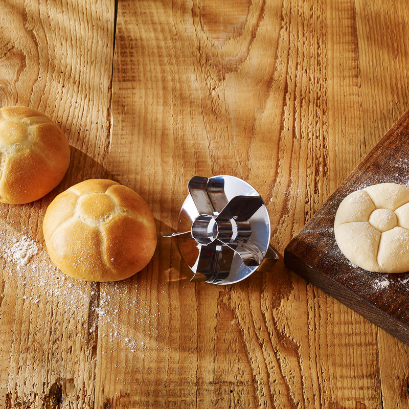 Klassischer Edelstahl-Brötchenstempel mit geformtem Teigling und frisch gebackenen Brötchen auf Holzoberfläche.
