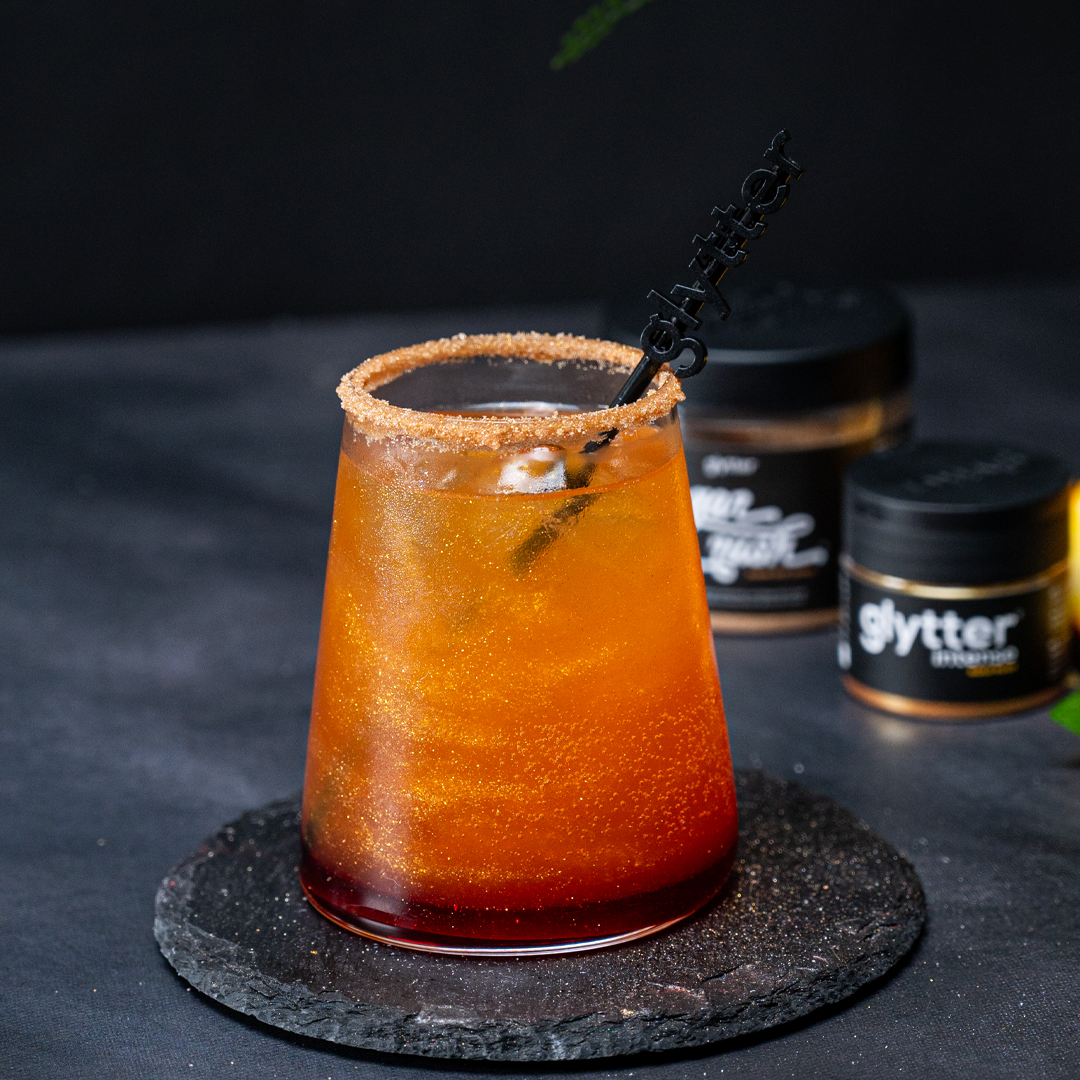Glytter Intense Gold Rush – glitzerndes Getränk im Glas mit Zuckerrand und Produktdosen im Hintergrund