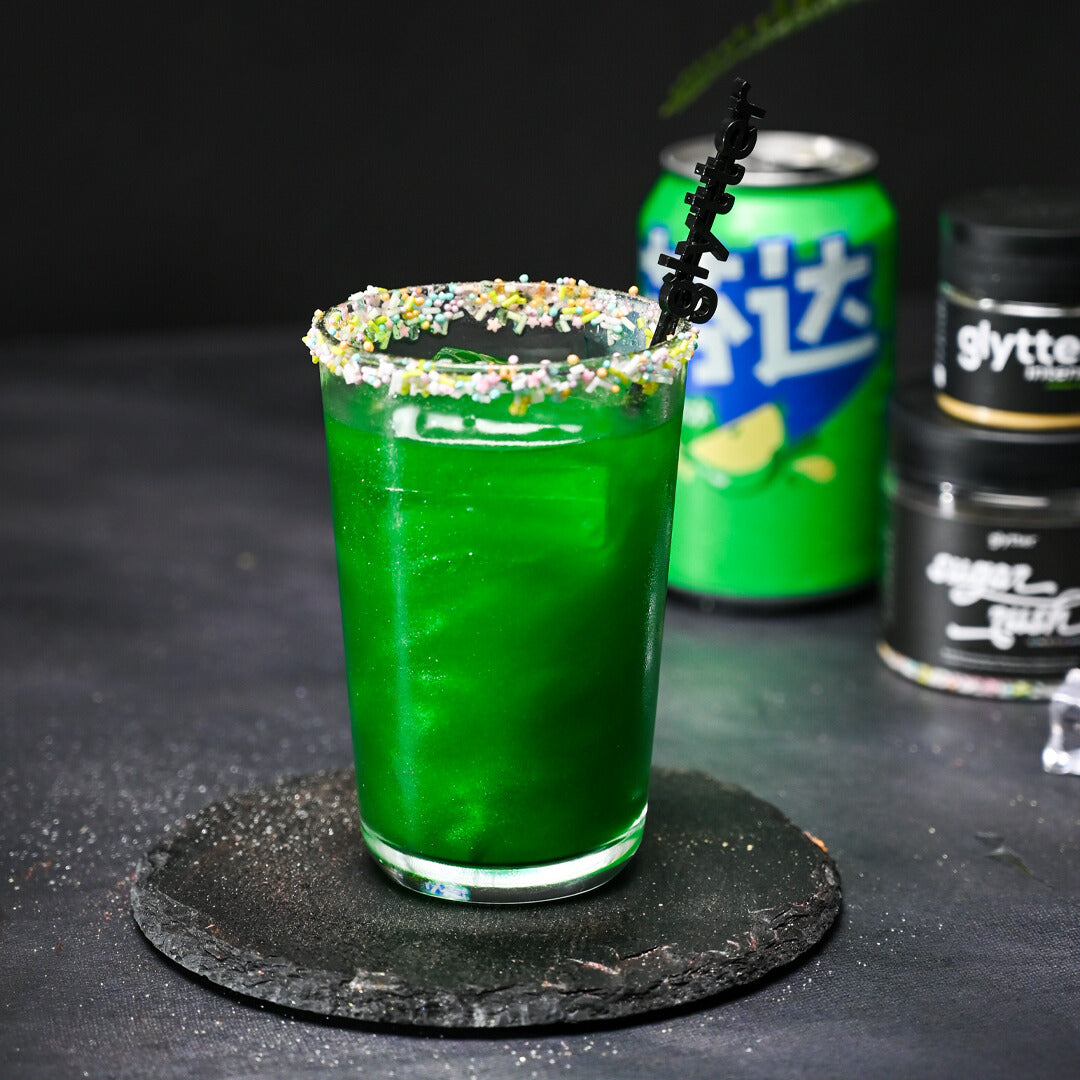 Decocino Glytter Intense Jungle Green – grünes glitzerndes Getränk im Glas mit buntem Zuckerrand und Produktdosen im Hintergrund