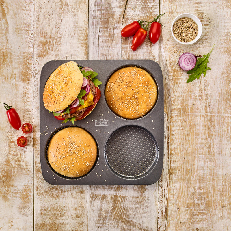 Perforierte Backform für Burger Buns mit frisch gebackenen Burgerbrötchen und einem fertig belegten Burger, umgeben von Tomaten, Zwiebeln und Sesam – Sallys, powered by Decocino
