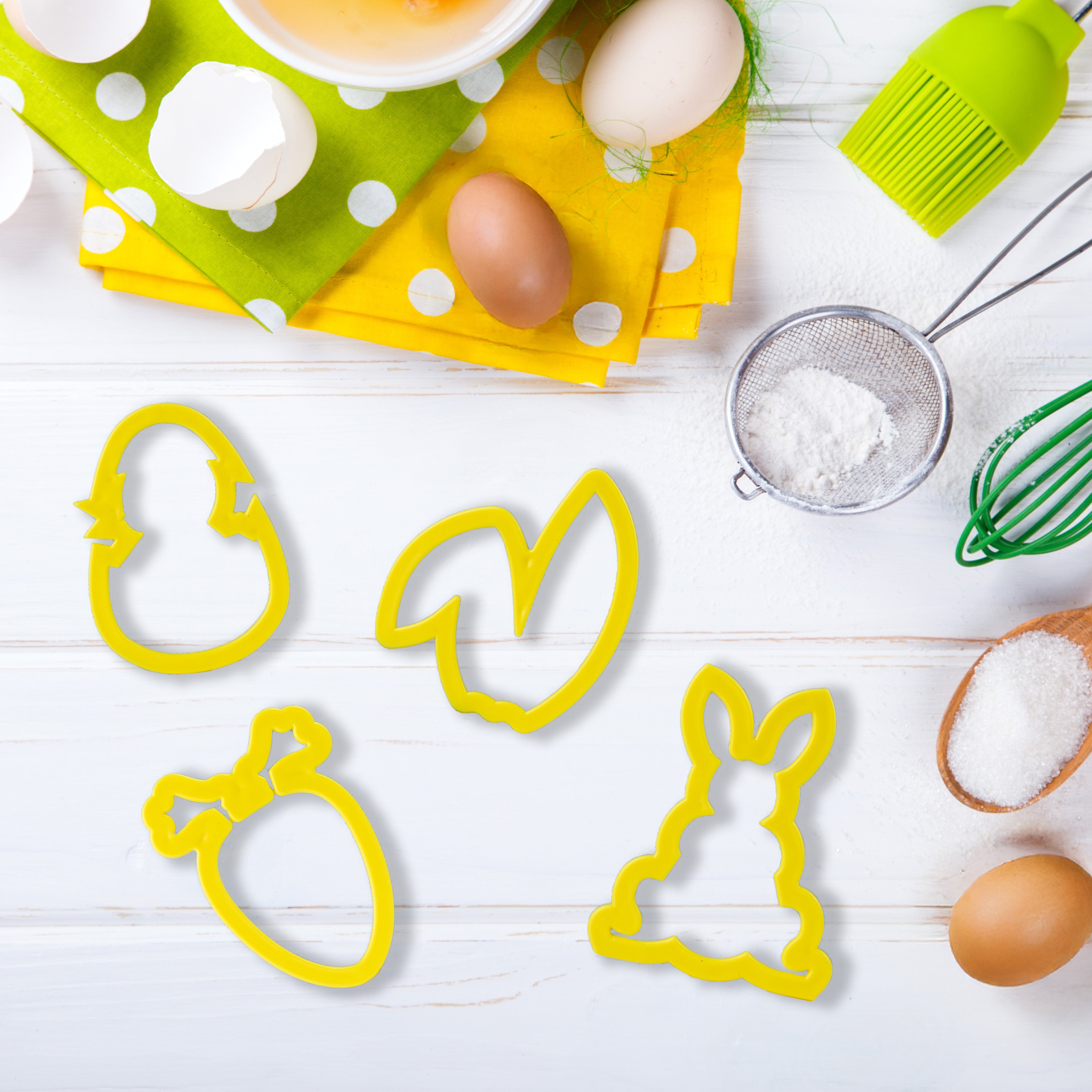 Gelbe Oster-Ausstechformen in Hase-, Ei-, Möhre- und Ohrenform auf einer weißen Arbeitsfläche, umgeben von Backzutaten